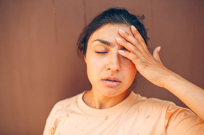 woman outside in the heat, sweating and holding her head with her eyes closed