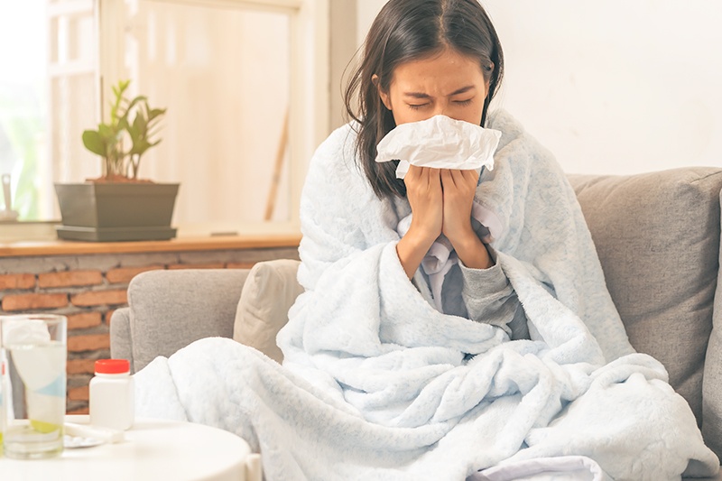 woman on couch wrapped in blanket blowing her nose. infection causing her to be sick