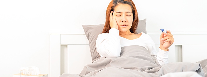 woman sick in bed holding her head and taking temperature