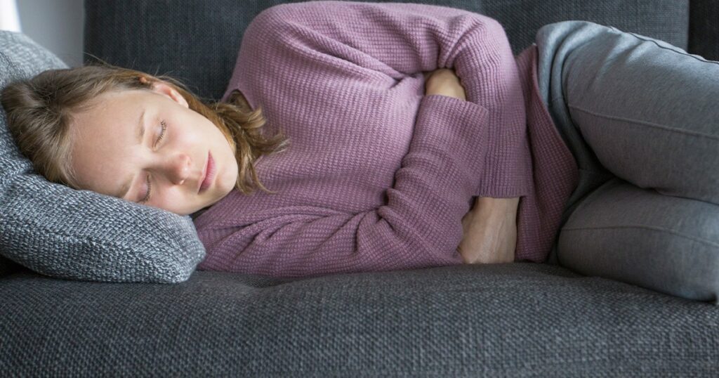 A young woman rests on a couch, holding her stomach in pain, showing signs of stomach flu.