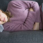 A young woman rests on a couch, holding her stomach in pain, showing signs of stomach flu.