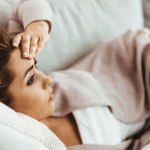 A woman reclines on a couch with one hand resting on her head and the other holding a thermometer as she checks her fever.