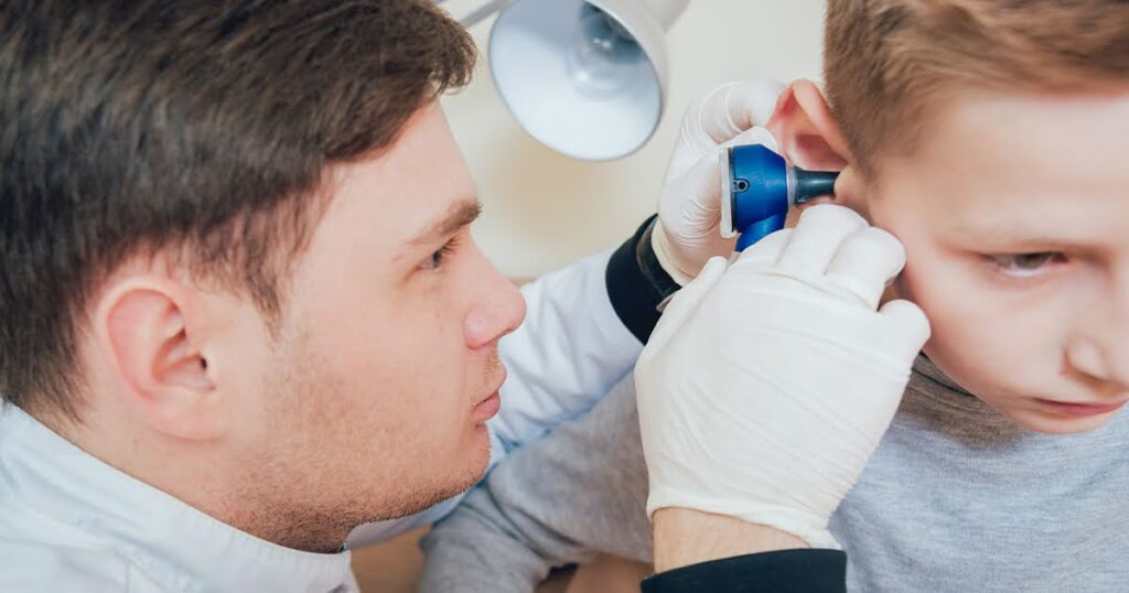A doctor examines a young boy's ears for fluid in the ear, with medical tools visible in the background.