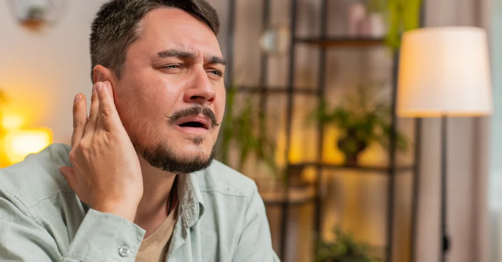 Man with a pained expression touching his ear with his hand, showing ear pain or discomfort.