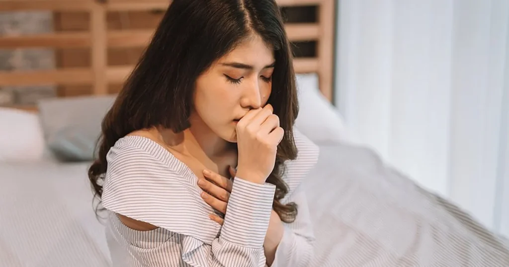 A close-up of a young woman sitting on a bed, leaning forward while coughing into her fist. She has her other hand placed over her heart in a gesture of discomfort. She is wearing a blue and white striped off-the-shoulder top in a neutrally decorated bedroom.
