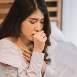 A close-up of a young woman sitting on a bed, leaning forward while coughing into her fist. She has her other hand placed over her heart in a gesture of discomfort. She is wearing a blue and white striped off-the-shoulder top in a neutrally decorated bedroom.
