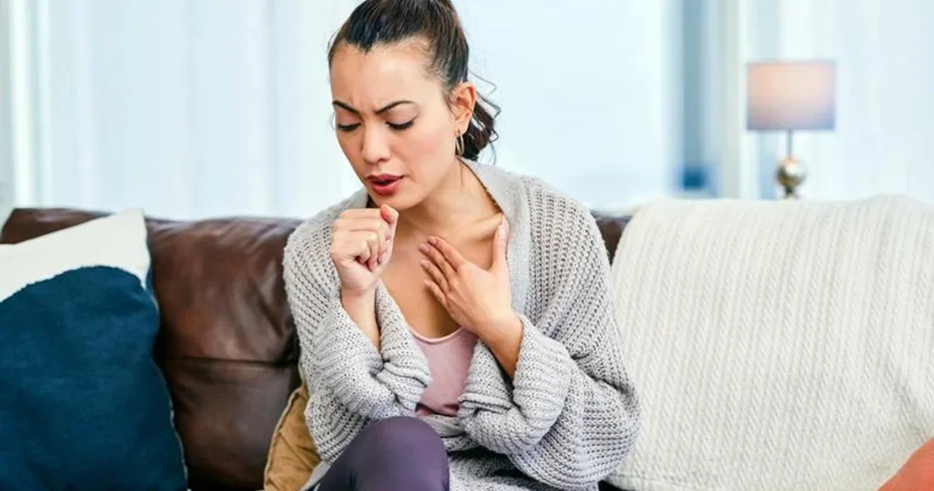 Woman sitting on a brown sofa in a bright living room, coughing into her hand while holding her chest, representing respiratory discomfort or persistent cough symptoms.