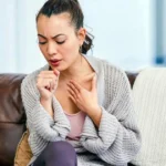 Woman sitting on a brown sofa in a bright living room, coughing into her hand while holding her chest, representing respiratory discomfort or persistent cough symptoms.