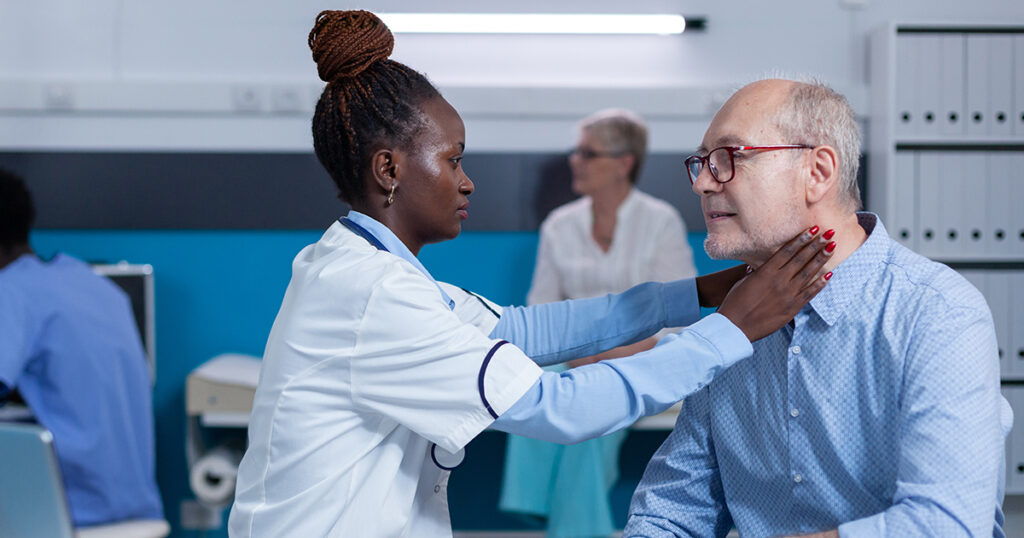 A doctor in a white coat examines an older man's neck and throat area in a medical office setting.