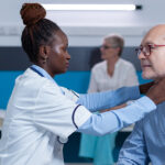 A doctor in a white coat examines an older man's neck and throat area in a medical office setting.