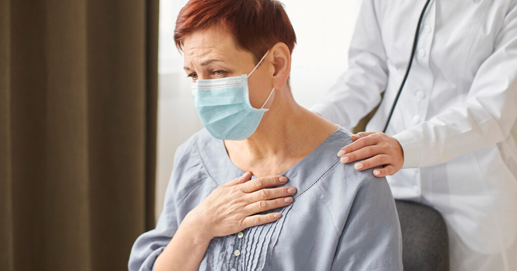 A woman wearing a face mask clutches her chest in discomfort as a medical professional offers support.
