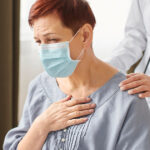 A woman wearing a face mask clutches her chest in discomfort as a medical professional offers support.