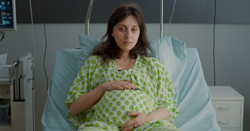 A pregnant woman in a hospital gown sits in bed, holding her stomach while looking tired or unwell.