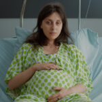 A pregnant woman in a hospital gown sits in bed, holding her stomach while looking tired or unwell.