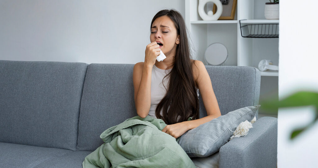 Sick woman covered in a blanket coughing on a sofa.