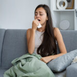 Sick woman covered in a blanket coughing on a sofa.