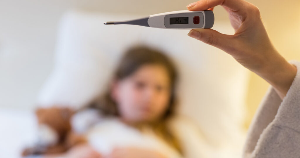 Hand holding a digital thermometer with a blurred child behind.