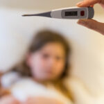 Hand holding a digital thermometer with a blurred child behind.