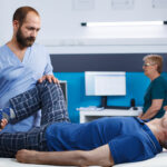 Physical therapist guiding an elderly man through a leg stretch.