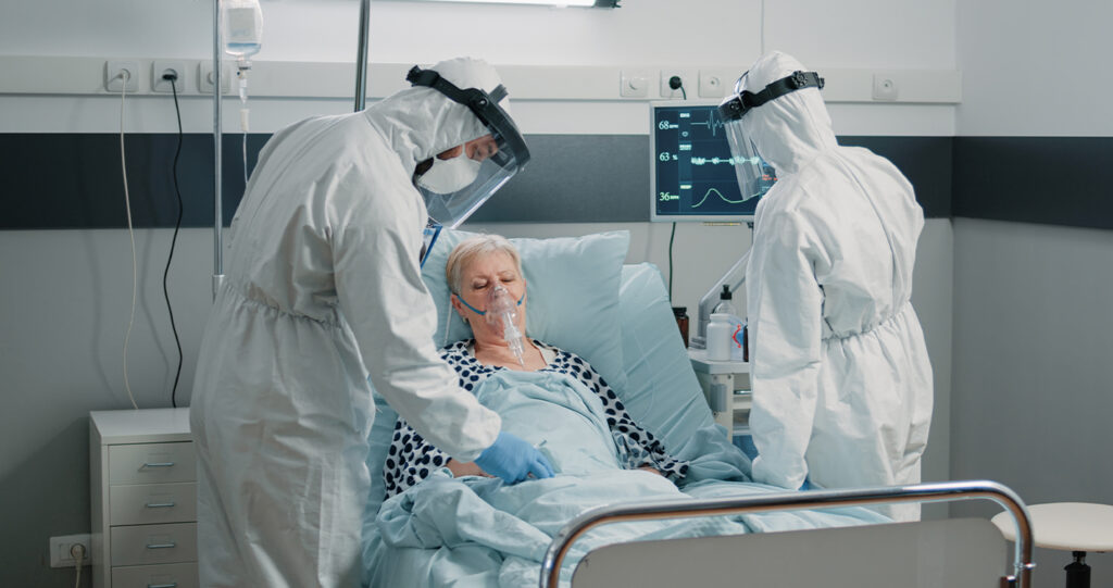 Medical staff in full protective gear treating an elderly patient wearing an oxygen mask.