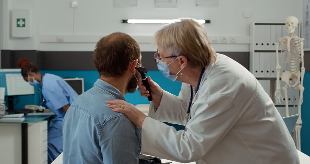 A female doctor wearing a face mask uses an otoscope to examine a male patient's ear in a medical office.