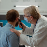 A female doctor wearing a face mask uses an otoscope to examine a male patient's ear in a medical office.
