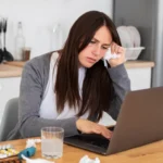 Fatigued woman working on a laptop surrounded by allergy medication.
