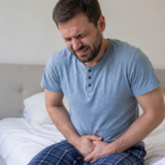 Man sitting on a bed holding his lower abdomen in pain.