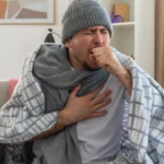 Man wrapped in blanket coughing while holding his aching chest.